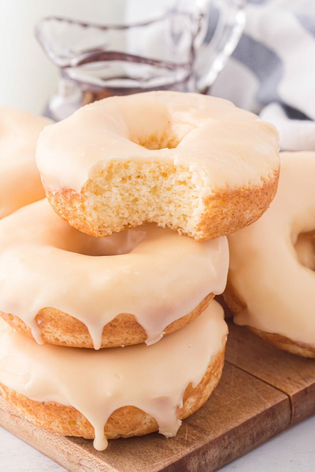 A stack of three maple-glazed donuts sits on a wooden board. The top donut has a large bite taken out, showing its soft, fluffy texture&mdash;a tempting scene for maple dessert recipes fans. In the background, there&rsquo;s syrup and a blurred striped cloth. // FoodNouveau.com