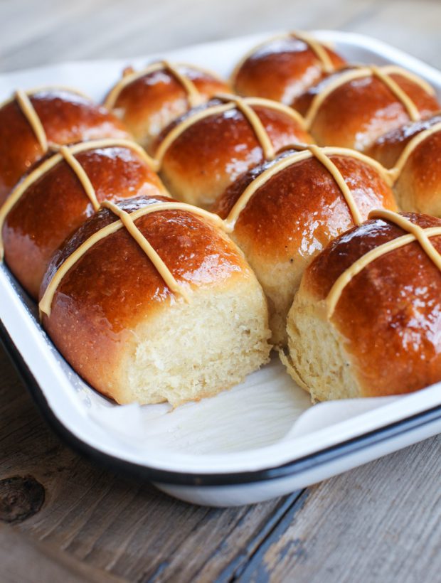 A tray of freshly baked hot cross buns, golden brown and shiny, with white icing crosses on top. One bun has been pulled apart, revealing the soft, fluffy inside&mdash;perfect inspiration for maple dessert recipes. The tray rests on a rustic wooden table. // FoodNouveau.com