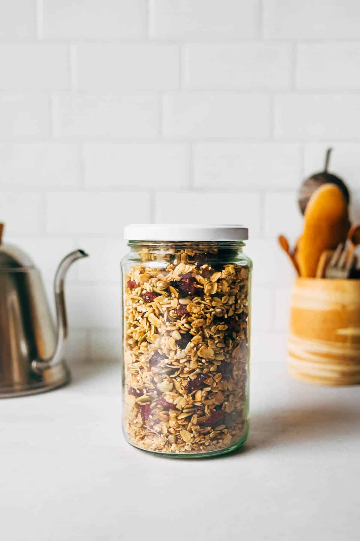 A glass jar filled with maple granola sits on a white countertop, perfect for maple dessert recipes. In the background, a silver teapot and wooden container of utensils rest against a white tiled wall. // FoodNouveau.com