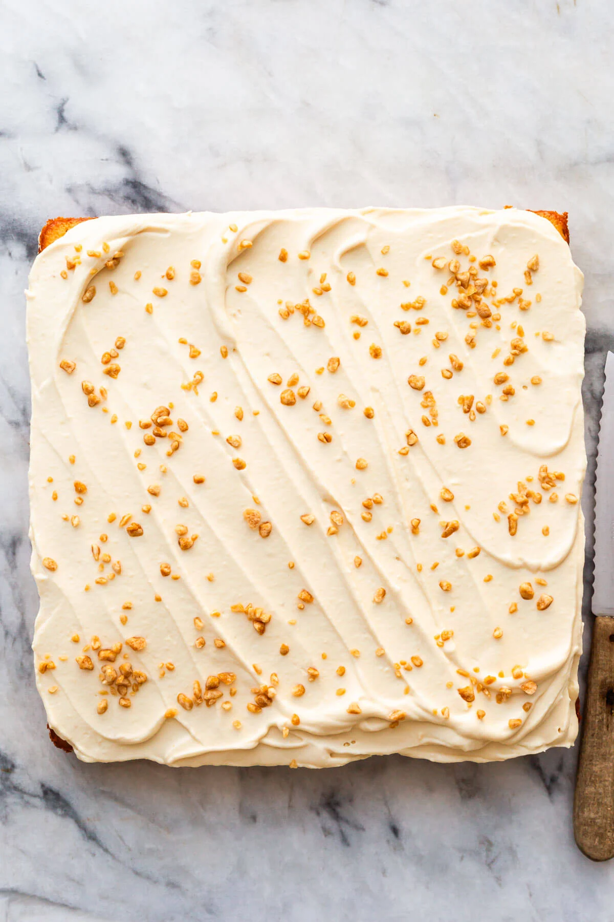 A square cake with smooth, maple whipped cream and sprinkled toffee bits on top sits on a marble surface next to a serrated knife with a wooden handle&mdash;perfect inspiration for maple dessert recipes. // FoodNouveau.com