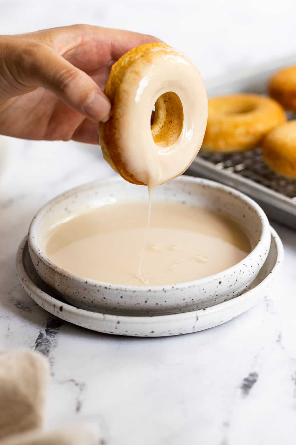 A hand dips a glazed donut into a bowl of maple glaze, with glaze dripping off the donut&mdash;perfect inspiration for maple dessert recipes. Additional donuts are visible on a cooling rack in the background. // FoodNouveau.com