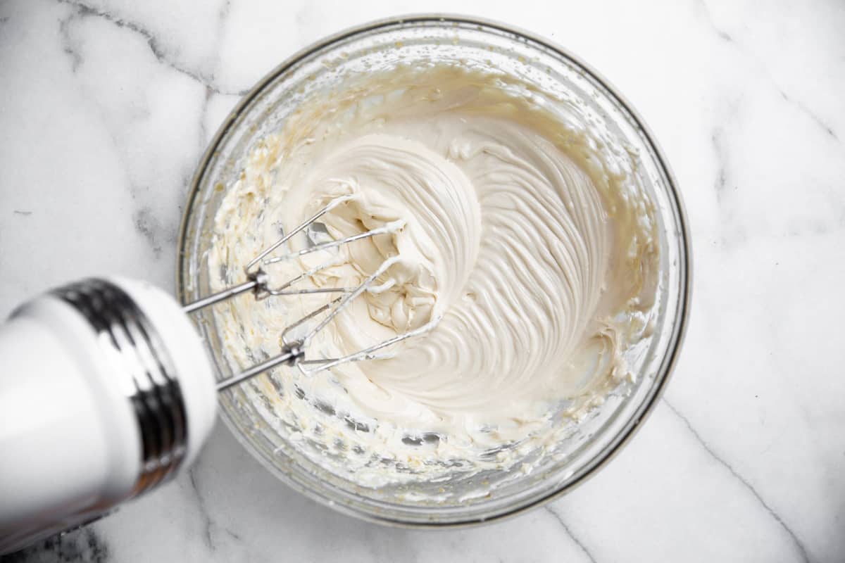 A glass bowl filled with creamy maple cream cheese frosting being blended by a hand mixer on a white marble surface&mdash;perfect for trying new maple dessert recipes. // FoodNouveau.com