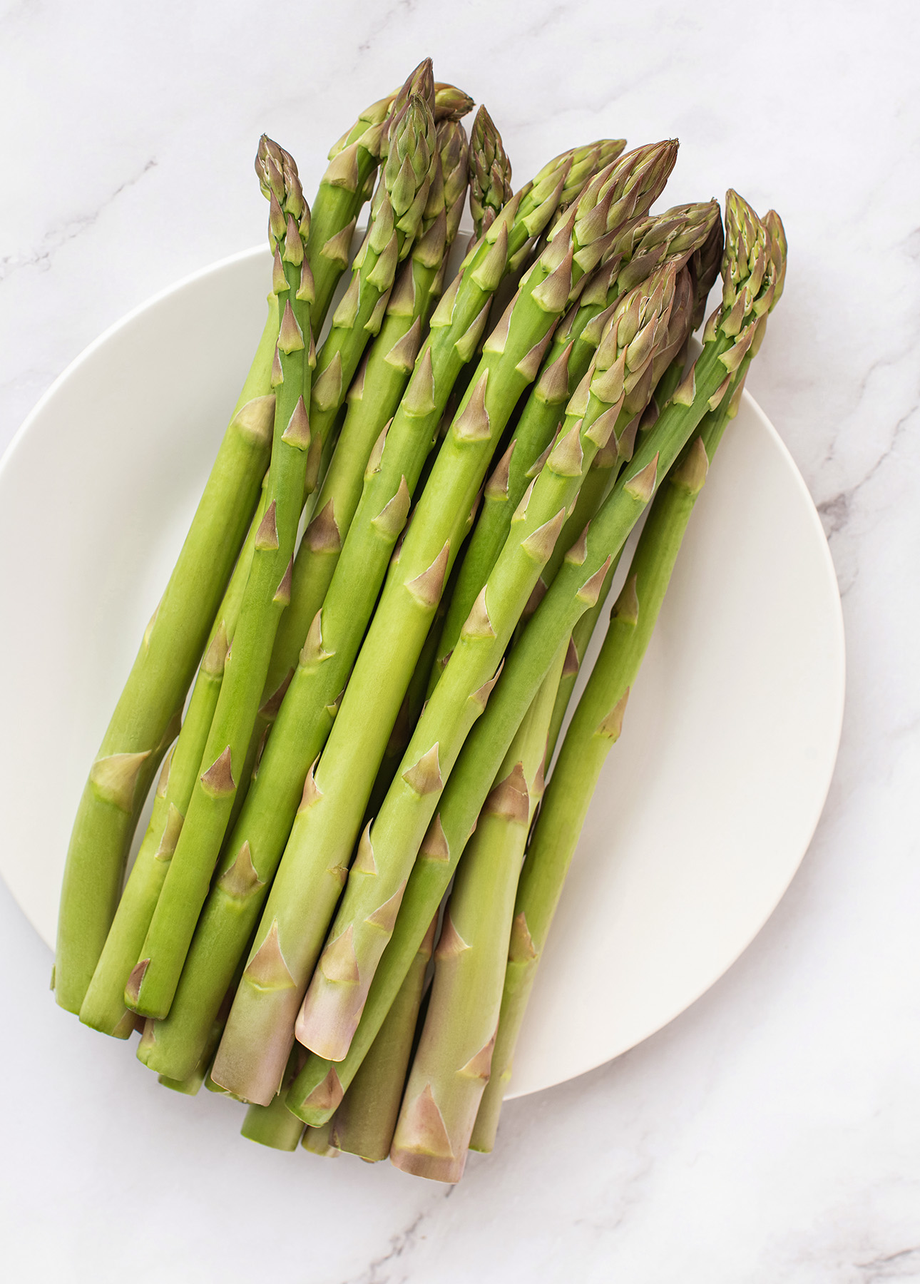 A bundle of fresh green asparagus spears arranged on a white plate, set on a light marble surface—perfect for preparing a vibrant lemon asparagus risotto. // FoodNouveau.com