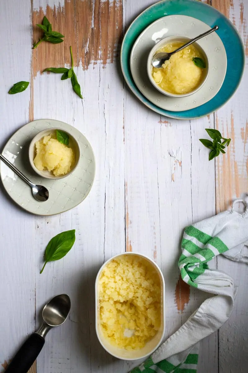 A container of yellow sorbet sits on a white wooden table, surrounded by two small bowls of sorbet, spoons, fresh basil leaves, a green-striped towel, and an ice cream scoop—perfect for fans of refreshing lemon dessert recipes. // FoodNouveau.com