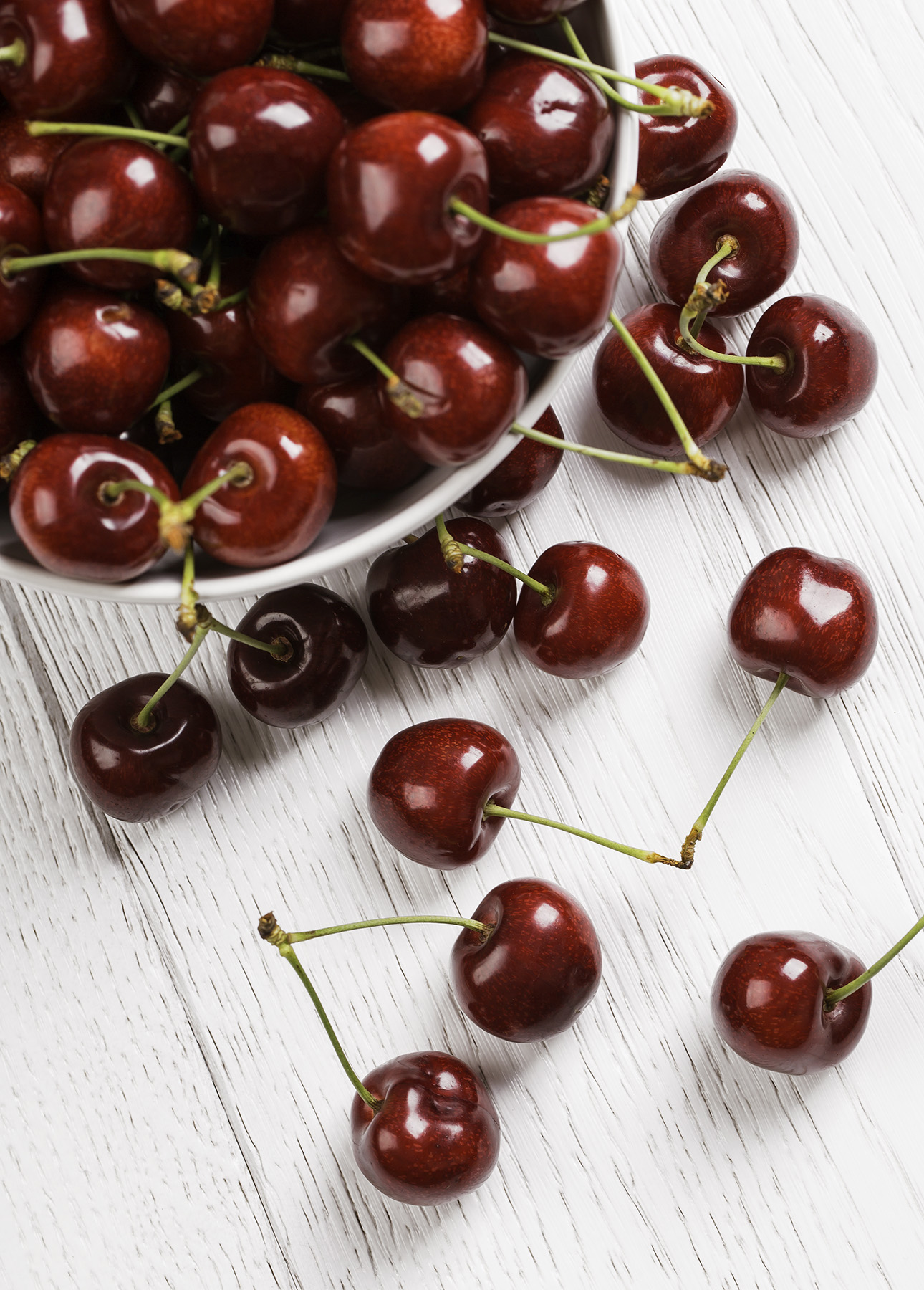 A white bowl filled with glossy, dark red cherries is tipped over on a white wooden surface, spilling some of the cherries onto the table. A few scattered cherries show off their vibrant green stems. // FoodNouveau.com