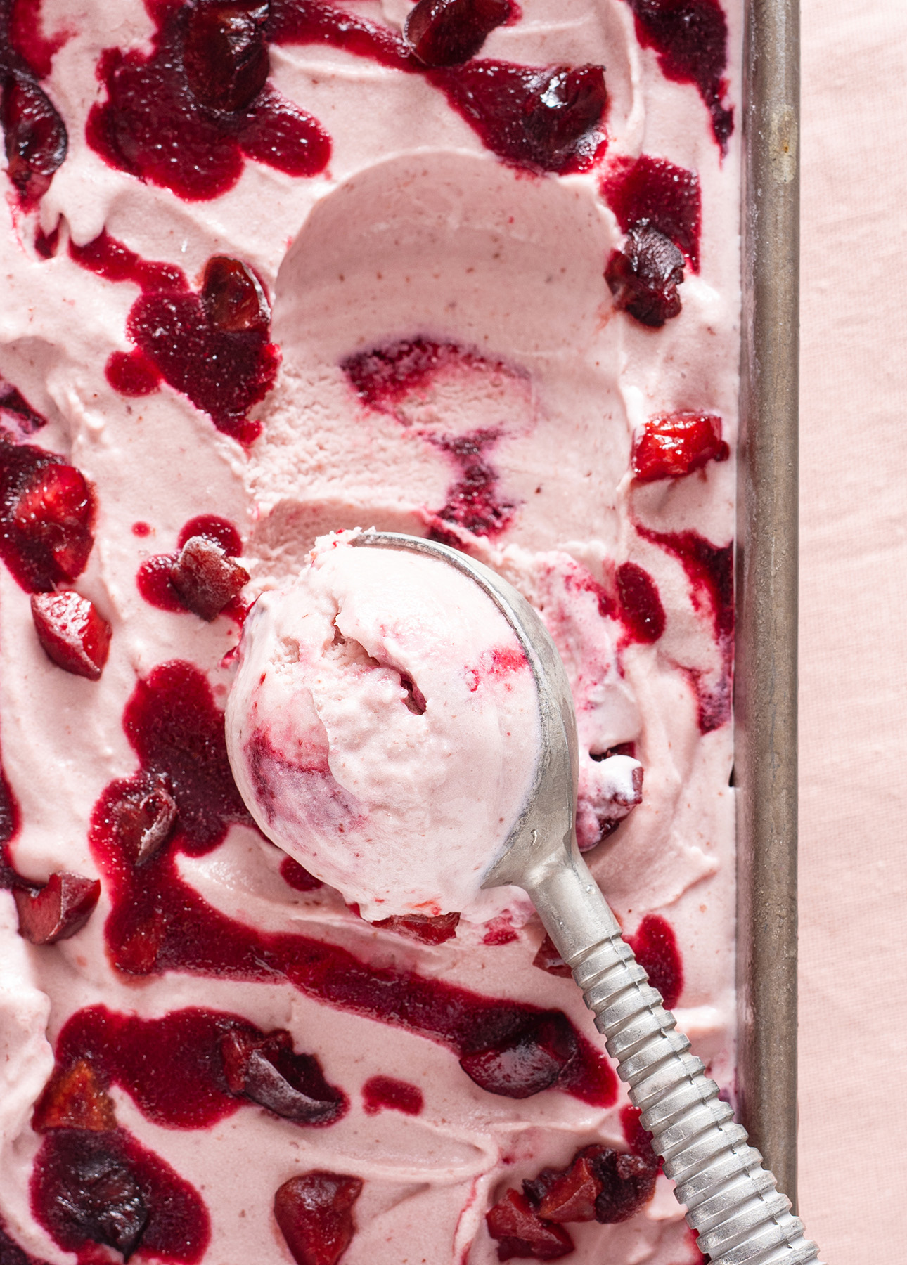 A metal ice cream scooper rests in a tray of pink Cherry Gelato. Dark red cherry puree swirls and scattered fruit pieces are visible on the surface. The smooth texture of the gelato contrasts with its vibrant toppings. // FoodNouveau.com