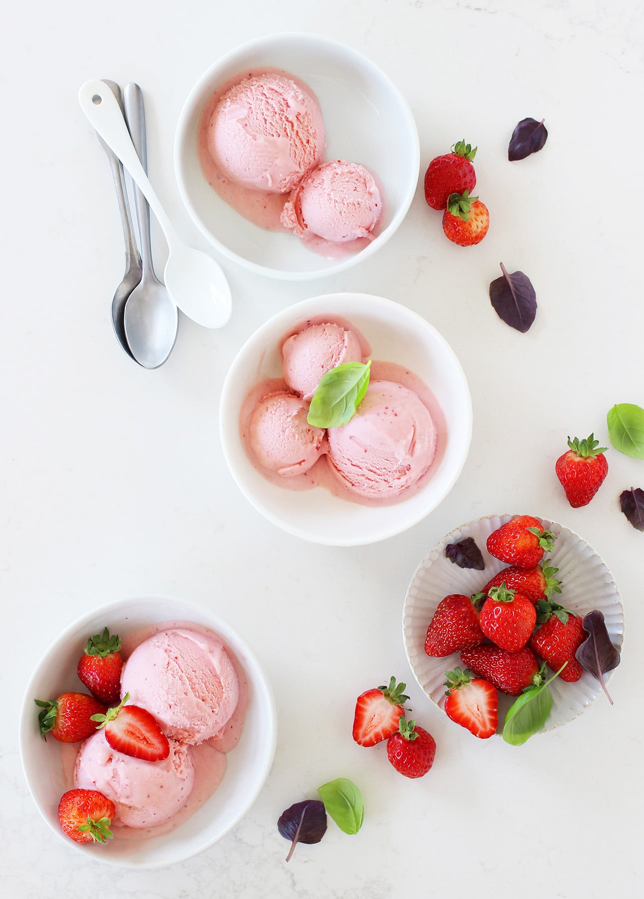 Three bowls of strawberry basil gelato, each topped with strawberries and basil leaves, sit on a white surface next to a small plate of fresh strawberries, loose leaves, and two spoons. // FoodNouveau.com