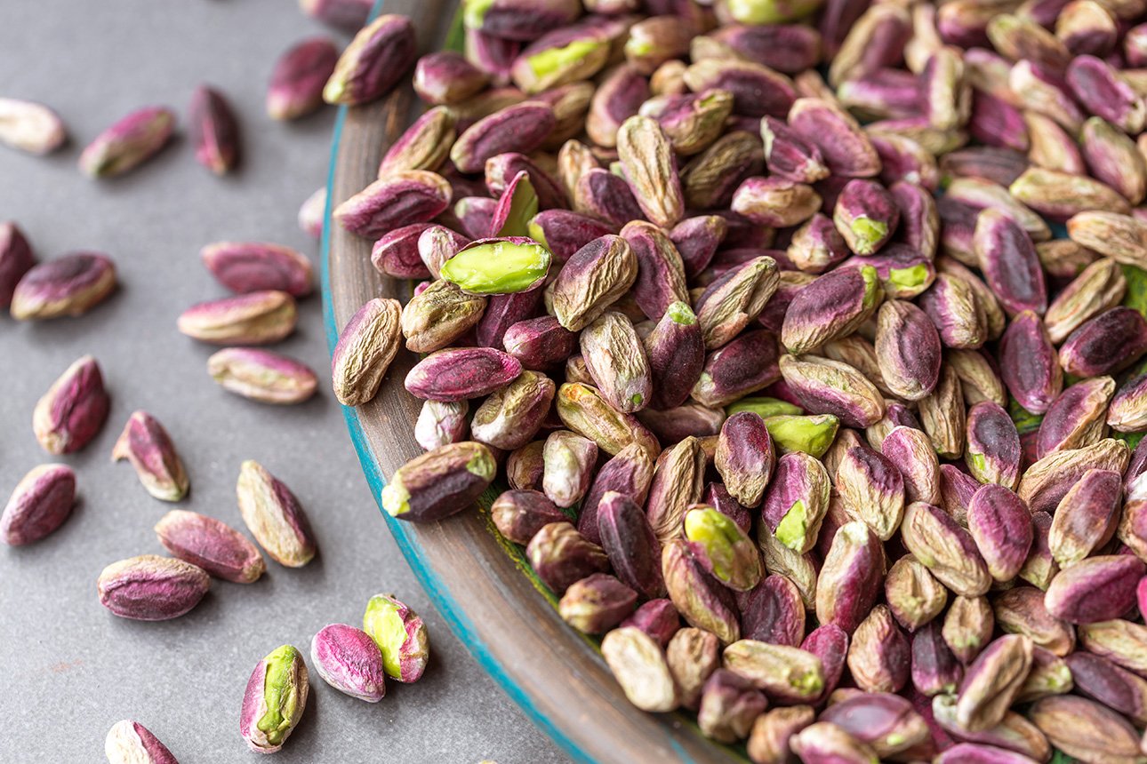 A close-up of a glass bowl filled with shelled, raw pistachios from Bronte, Sicily, some with purple and green hues, with a few pistachios scattered on a gray surface beside the bowl. // FoodNouveau.com