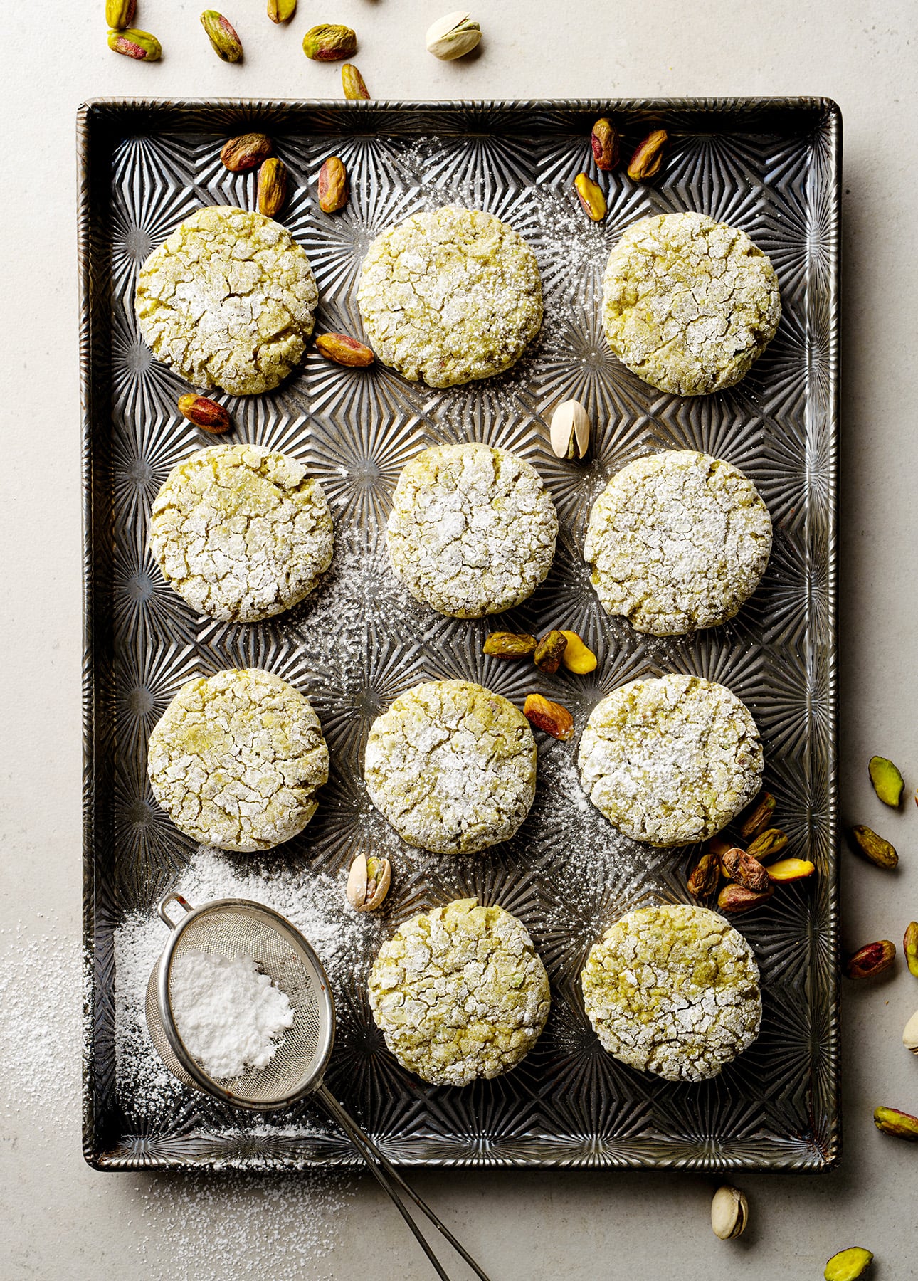 A baking tray with twelve round Italian pistachio cookies dusted with powdered sugar, surrounded by scattered whole pistachios. A metal sieve with powdered sugar sits in the corner of the tray. // FoodNouveau.com
