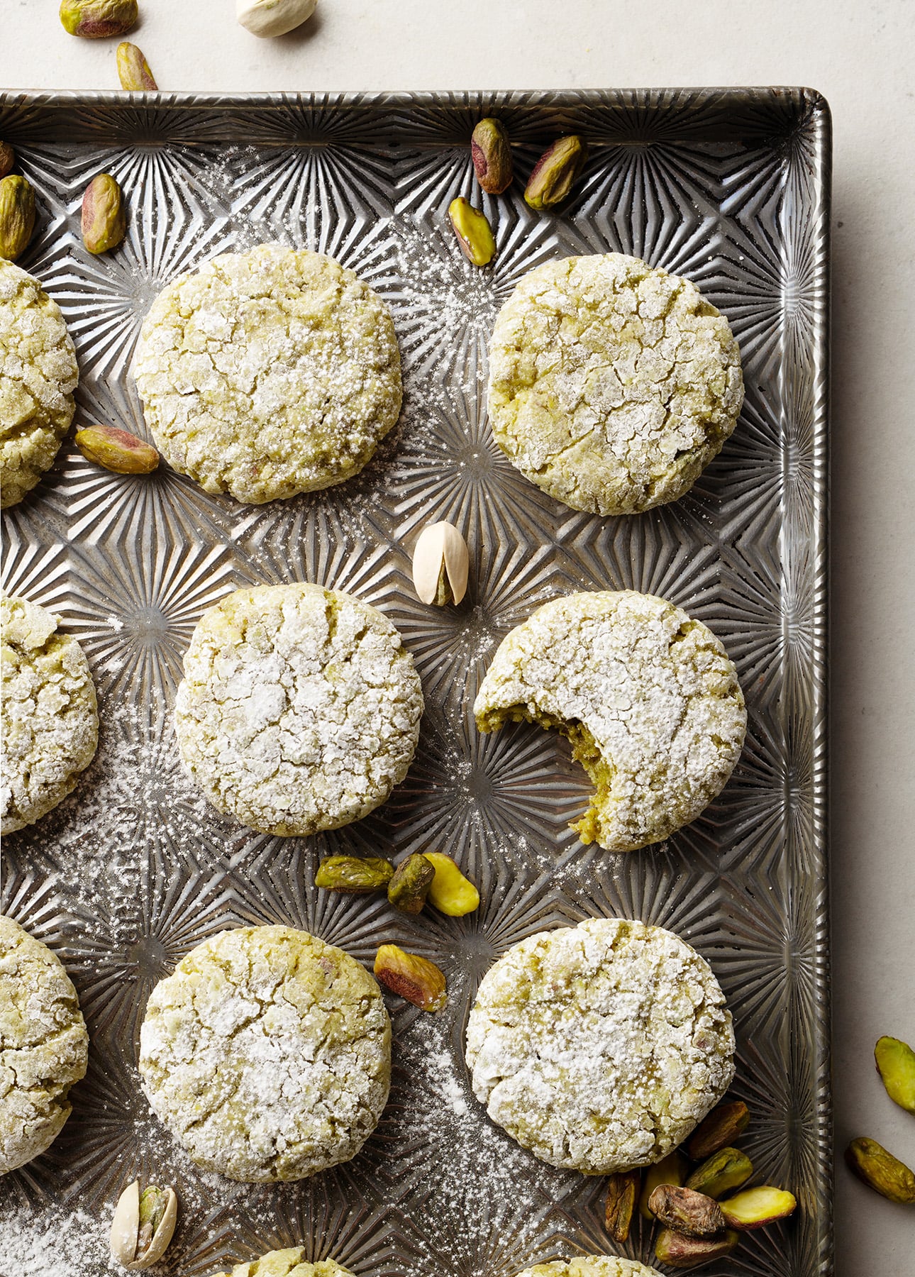 A baking sheet with round, powdered sugar-dusted pistachio cookies, some whole and one with a bite taken out, surrounded by scattered pistachio nuts. // FoodNouveau.com
