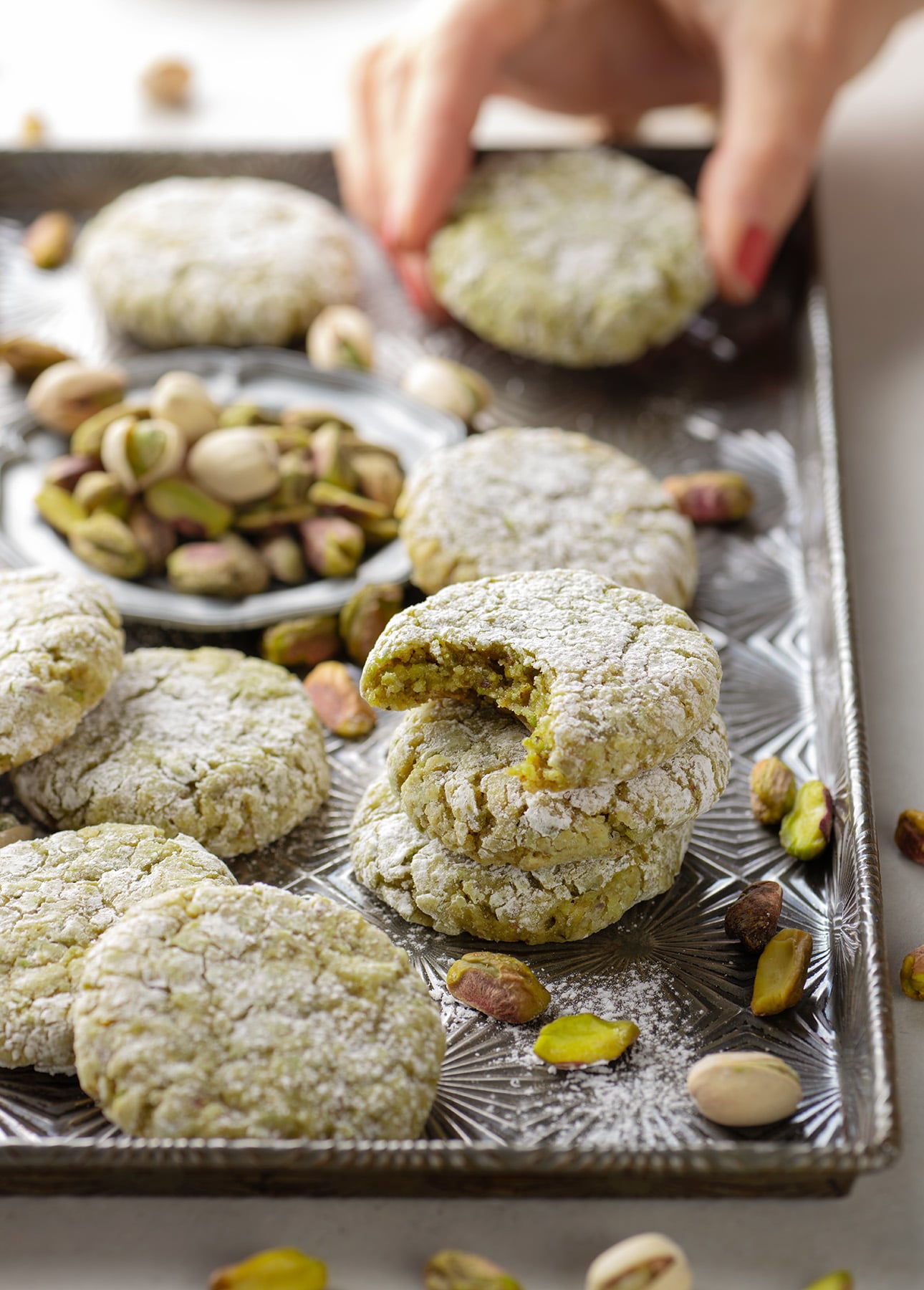 A tray of green pistachio cookies, some dusted with powdered sugar, with whole pistachios scattered around. One cookie has a bite taken out, and a hand is reaching for another. // FoodNouveau.com