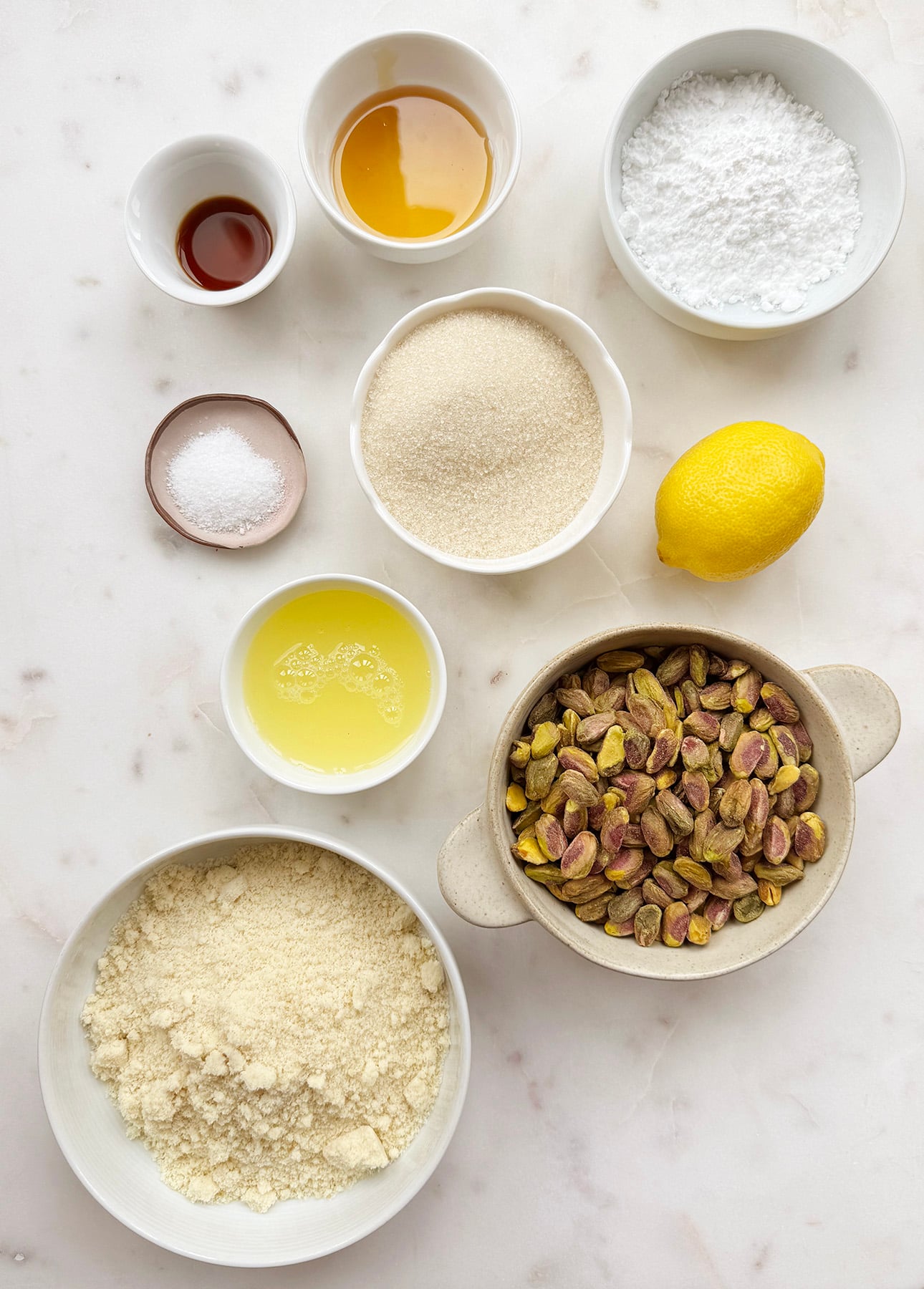 Bowls of ingredients to make Italian pistachio cookies on a white surface, including whole pistachios, almond flour, granulated sugar, powdered sugar, egg white, salt, vanilla extract, honey, and a whole lemon. // FoodNouveau.com