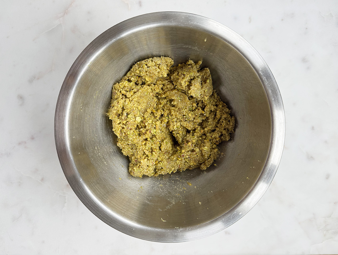 A stainless steel mixing bowl containing a coarse, yellow-green dough that will be used to make Italian pistachio cookies. The bowl sits on a white marble surface. // FoodNouveau.com