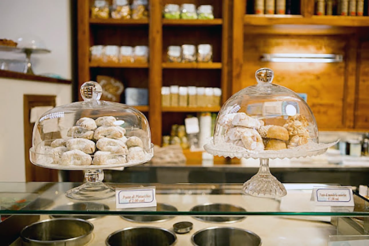 Two trays of assorted cookies are displayed under glass domes on an Italian bakery counter, with wooden shelves and jars in the background. Small signs with labels are placed in front of each tray. // FoodNouveau.com