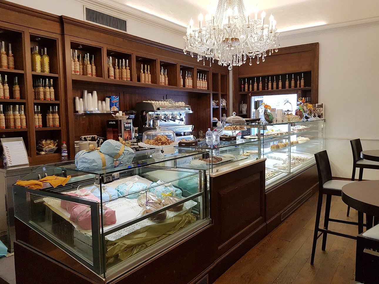 An Italian bakery with glass display cases filled with pastries and cakes, wooden shelves stocked with bottles and jars, a chandelier hanging from the ceiling, and tables and chairs for customers. // FoodNouveau.com