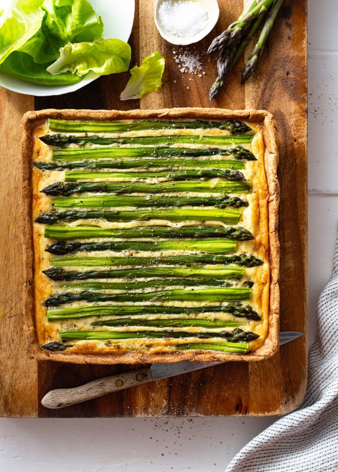 A rectangular asparagus tart sits on a wooden cutting board, topped with whole asparagus spears. A knife, a bowl of salt, loose lettuce leaves, and some raw asparagus are nearby. // FoodNouveau.com