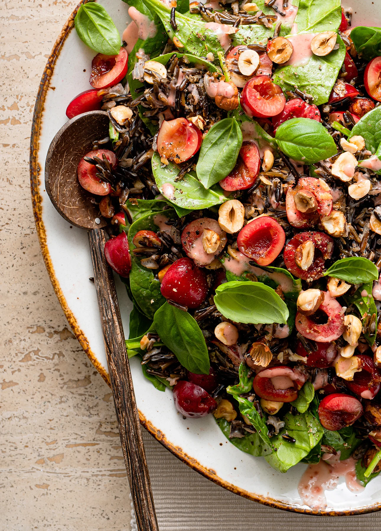 A salad with spinach, wild rice, cherries, toasted hazelnuts, and fresh basil on a white plate with a wooden spoon. Light pink dressing is drizzled on top. // FoodNouveau.com