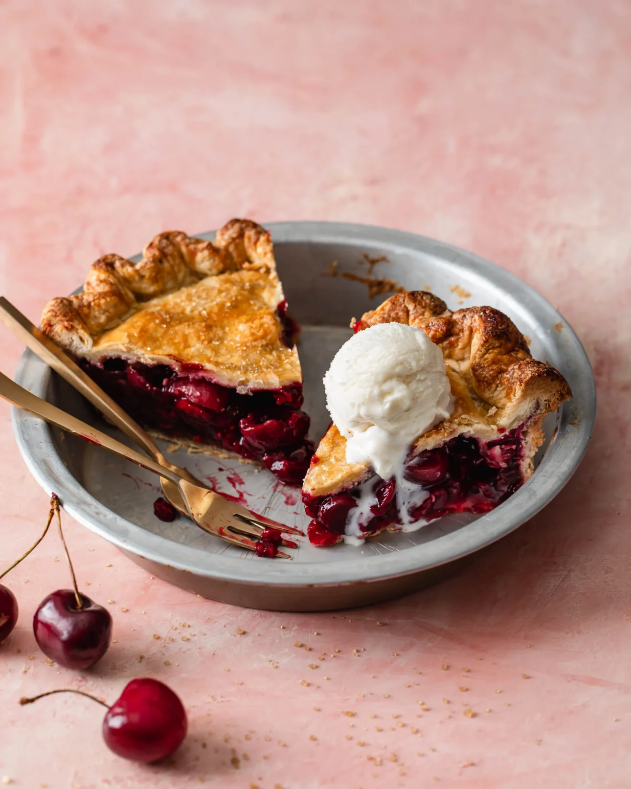 A metal pie pan holds two slices of cherry pie, one topped with vanilla ice cream. Two gold forks rest on the pan, and a few cherries are on the pink surface nearby. // FoodNouveau.com