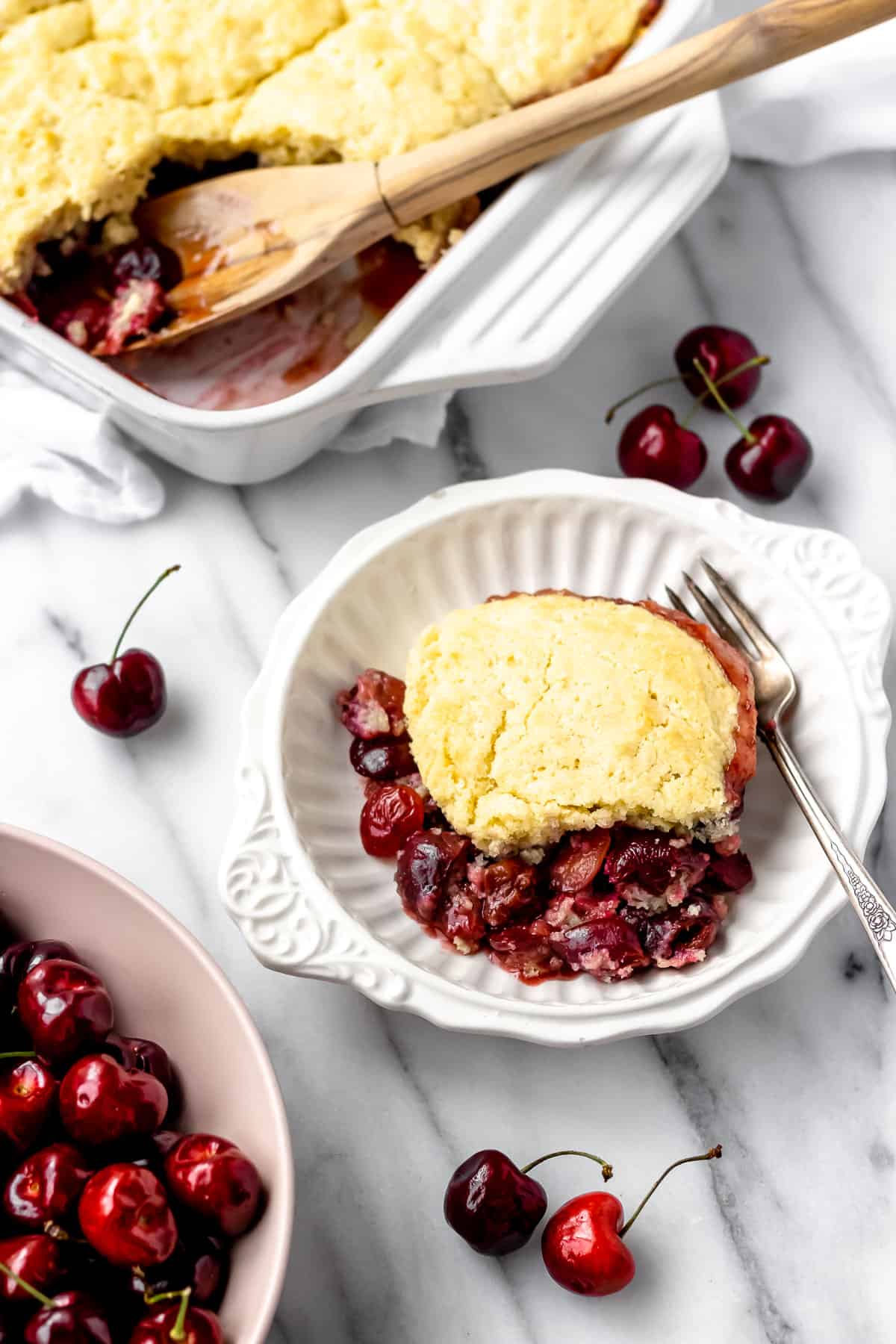 A serving of cherry cobbler on a white plate with a fork. Fresh cherries are scattered around, and a baking dish with more cobbler is in the background. // FoodNouveau.com