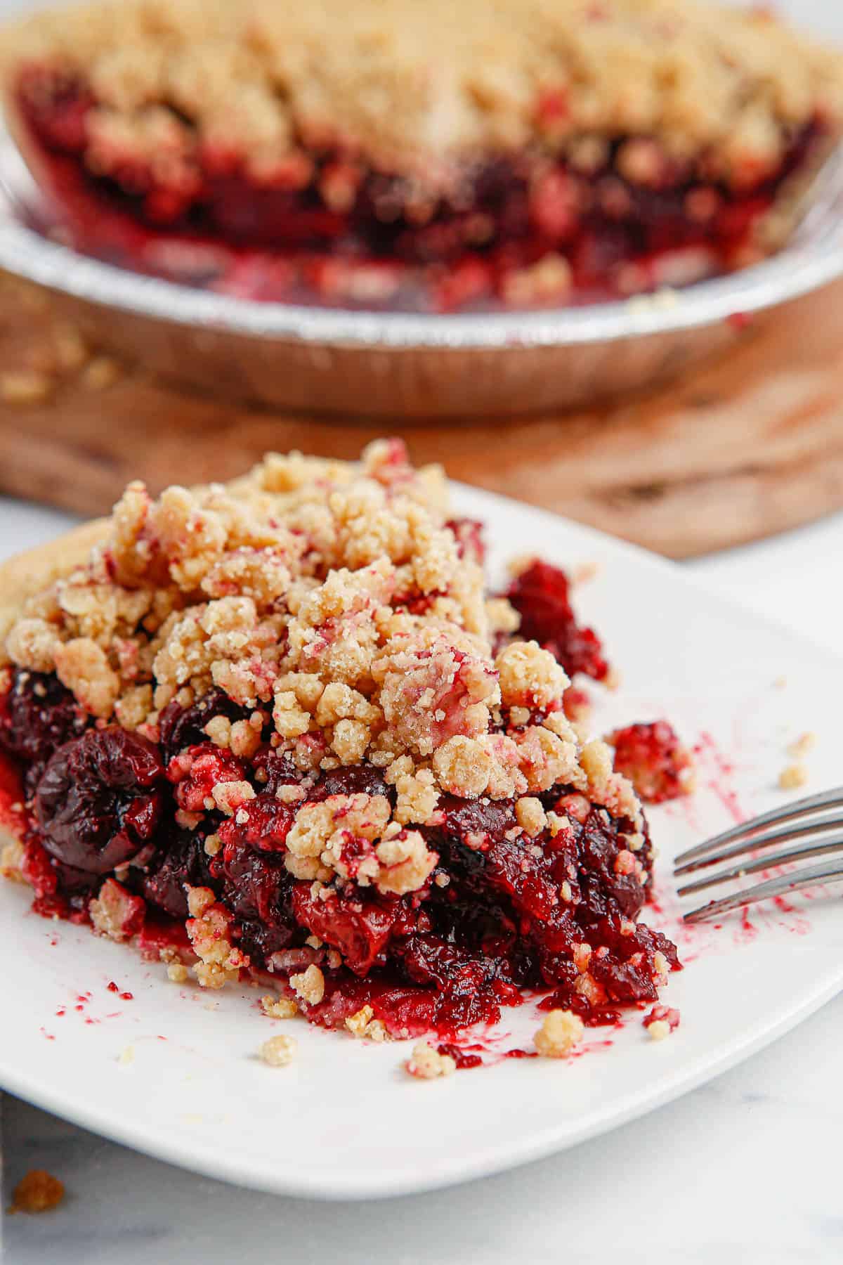 A slice of berry crumble pie with a buttery crumble topping sits on a white plate, with a fork beside it. The rest of the pie is in the background. // FoodNouveau.com