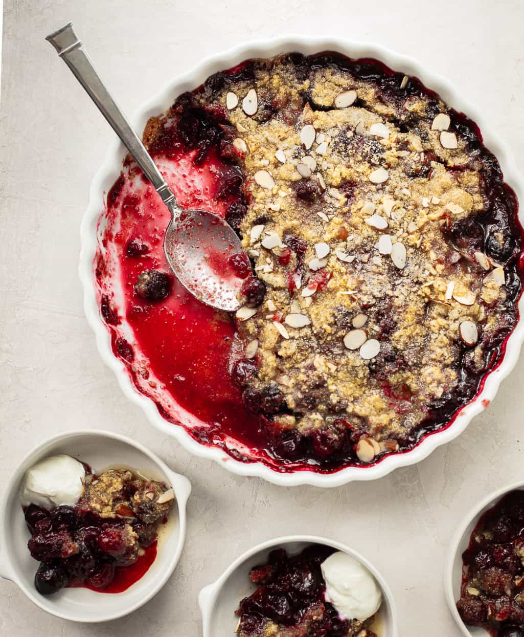 A round berry crumble in a white dish with a spoon, topped with sliced almonds. Three small bowls of crumble and whipped cream are served alongside. // FoodNouveau.com