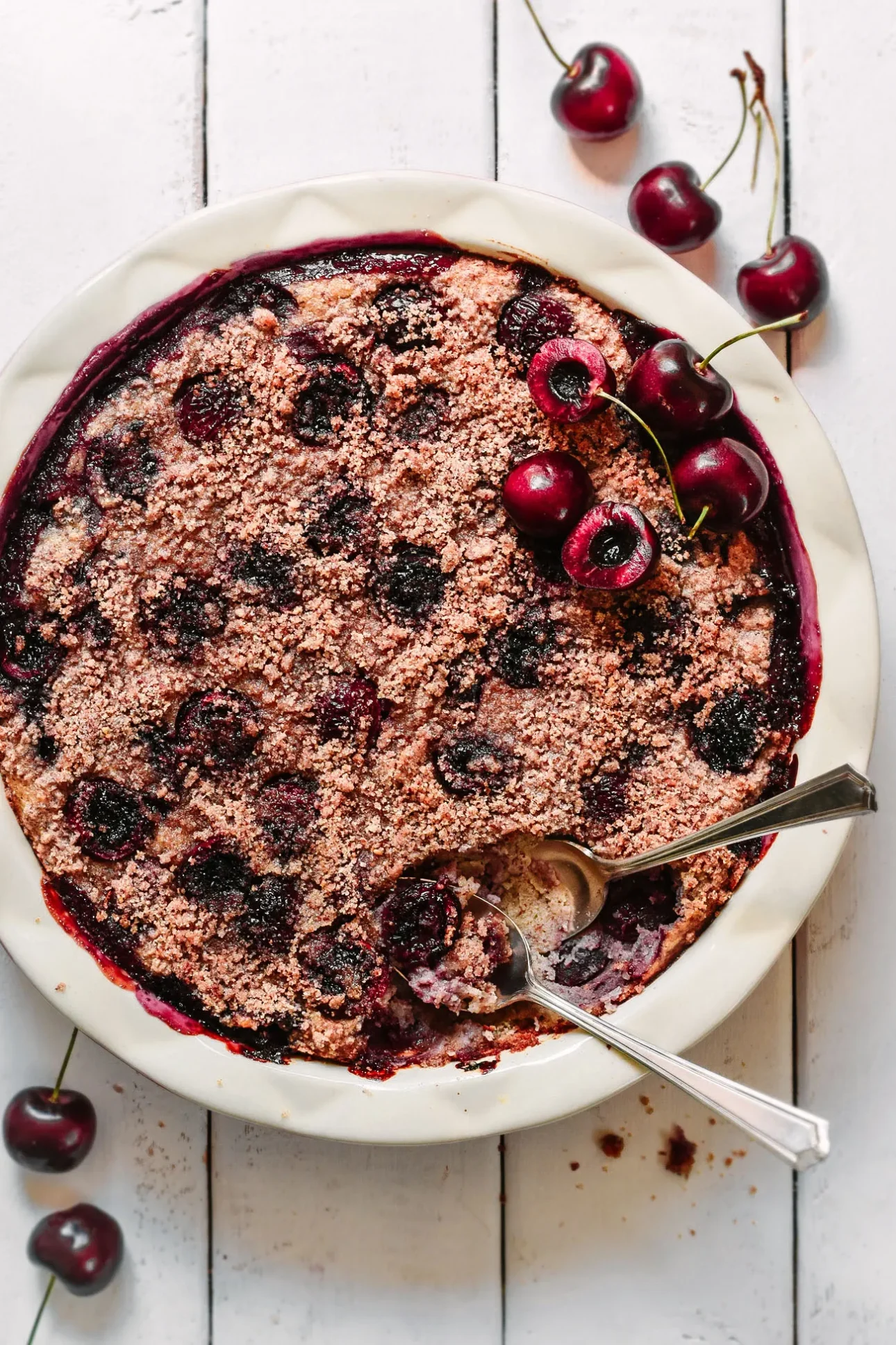 A cherry crumble dessert in a white dish with a serving spoon, partially scooped out. Fresh cherries, some whole and some halved, are scattered on top and around the dish on a white wooden surface. // FoodNouveau.com