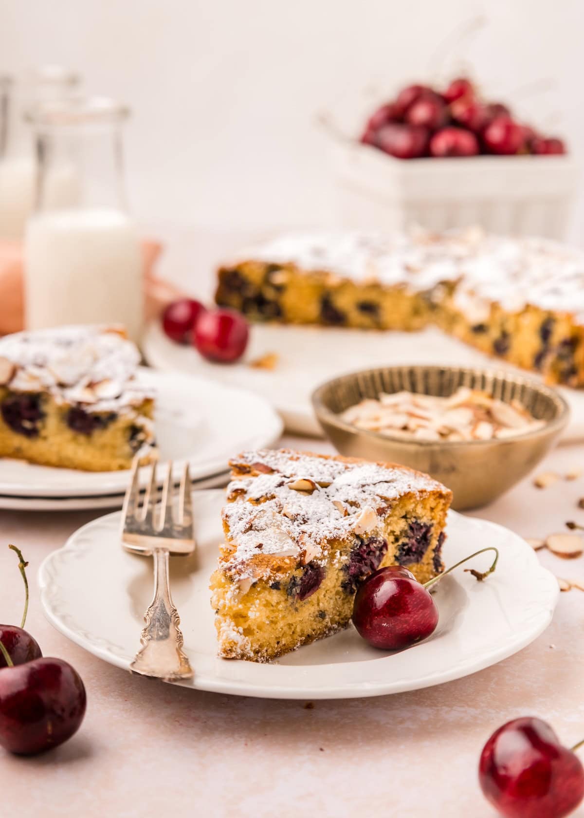 A slice of cherry almond cake on a white plate with fresh cherries, a fork, and bowls of sliced almonds and more cake in the background. A milk bottle and a basket of cherries are also visible. // FoodNouveau.com