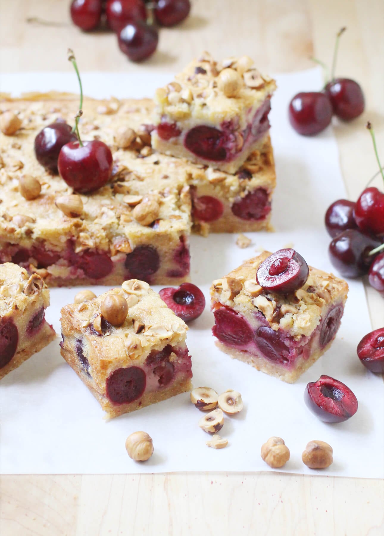 Cherry dessert bars with chopped hazelnuts on top, sliced into squares and arranged on a light surface, surrounded by whole and halved cherries and scattered hazelnuts. // FoodNouveau.com