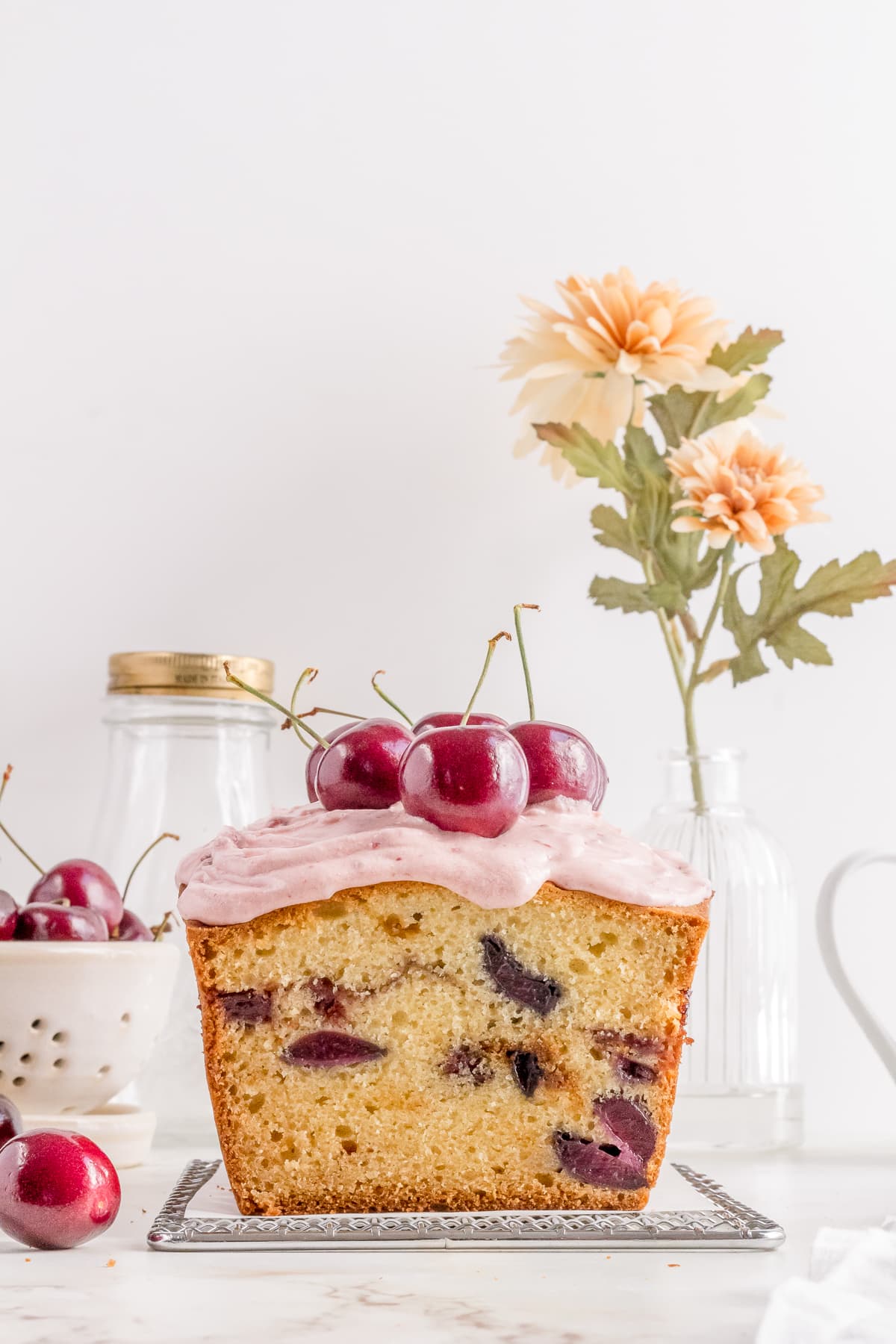 A loaf cake with pink frosting and cherries on top sits on a cooling rack, with a jar, colander of cherries, and a vase of flowers in the background. // FoodNouveau.com