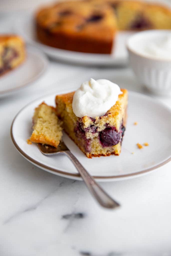 A slice of cherry cake topped with whipped cream sits on a white plate with a fork, with more cake and a cup of cream in the background. // FoodNouveau.com