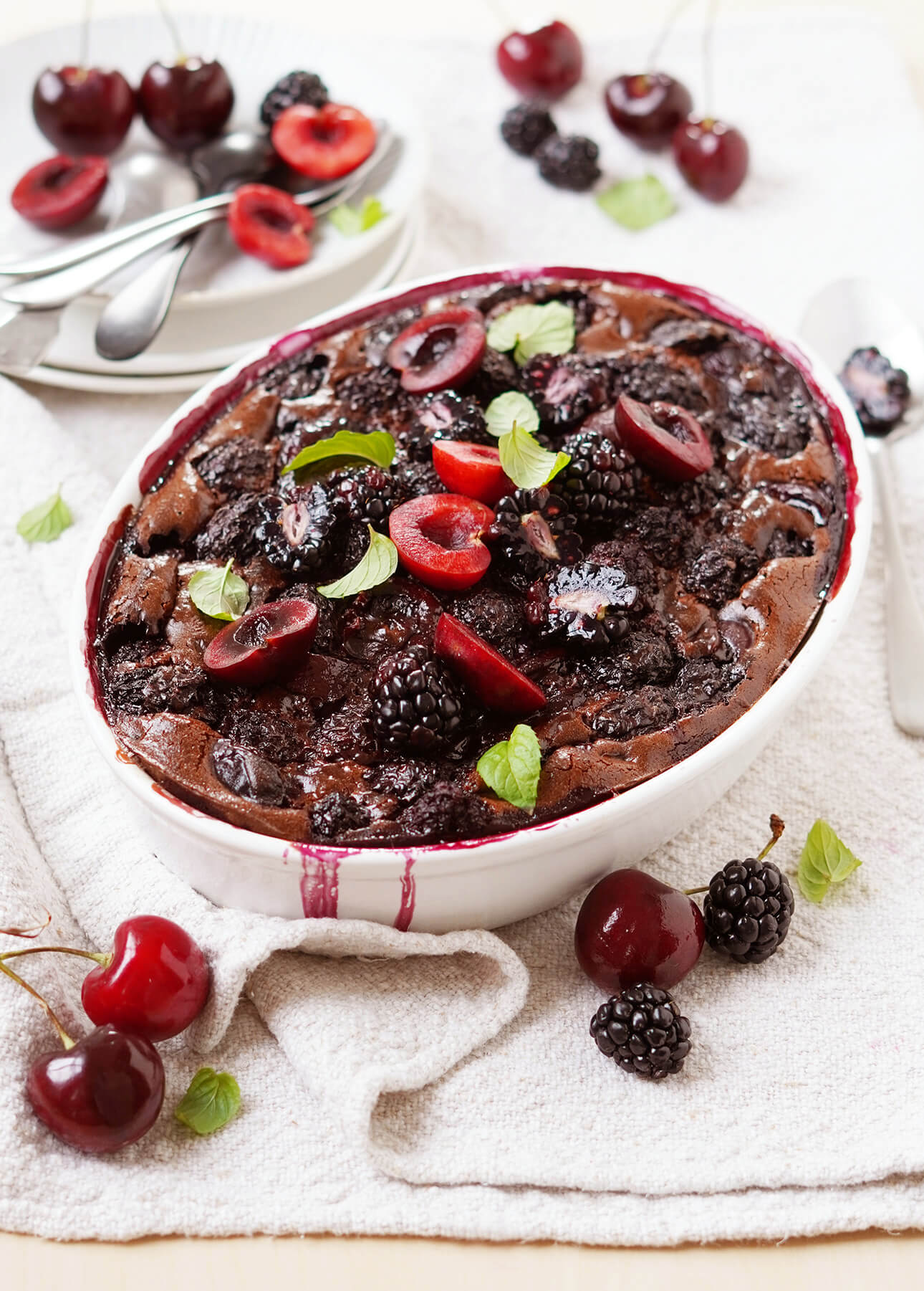 A baked chocolate dessert topped with fresh blackberries, cherries, and mint leaves sits on a white towel, with more berries and cherries scattered around. Plates and utensils are in the background. // FoodNouveau.com