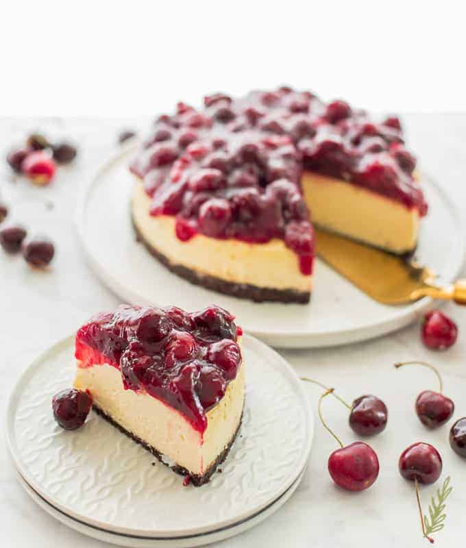 A slice of cherry-topped cheesecake on a plate, with the rest of the cheesecake and scattered fresh cherries in the background. // FoodNouveau.com
