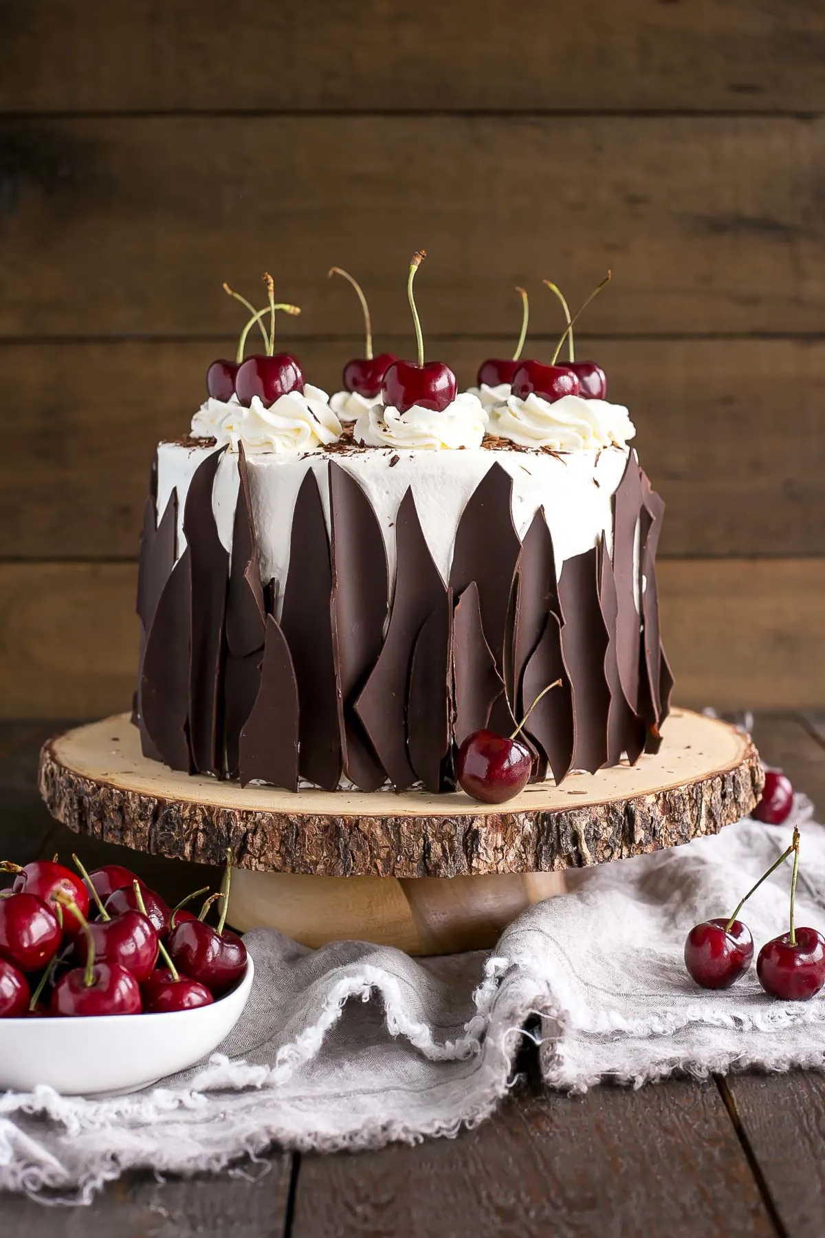 A Black Forest cake with whipped cream, dark chocolate shards, and cherries on top sits on a rustic wooden cake stand. A bowl of fresh cherries is beside it on a cloth. // FoodNouveau.com