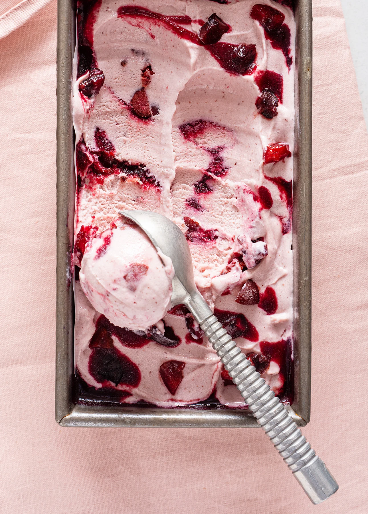 Rectangular metal container filled with pink ice cream swirled with dark red fruit sauce. A metal ice cream scoop rests on top. The container sits on a light pink cloth. // FoodNouveau.com