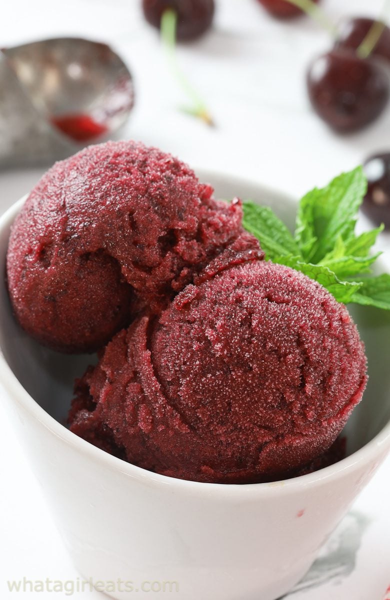 Two scoops of dark red cherry sorbet in a white bowl, garnished with a sprig of fresh mint. Cherries and an ice cream scoop are blurred in the background. // FoodNouveau.com