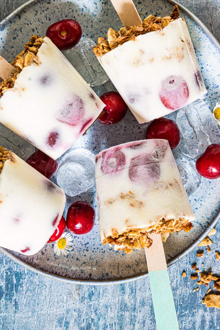 Four yogurt popsicles with granola and cherries on a ceramic plate, surrounded by fresh cherries and ice cubes, on a blue textured surface. // FoodNouveau.com