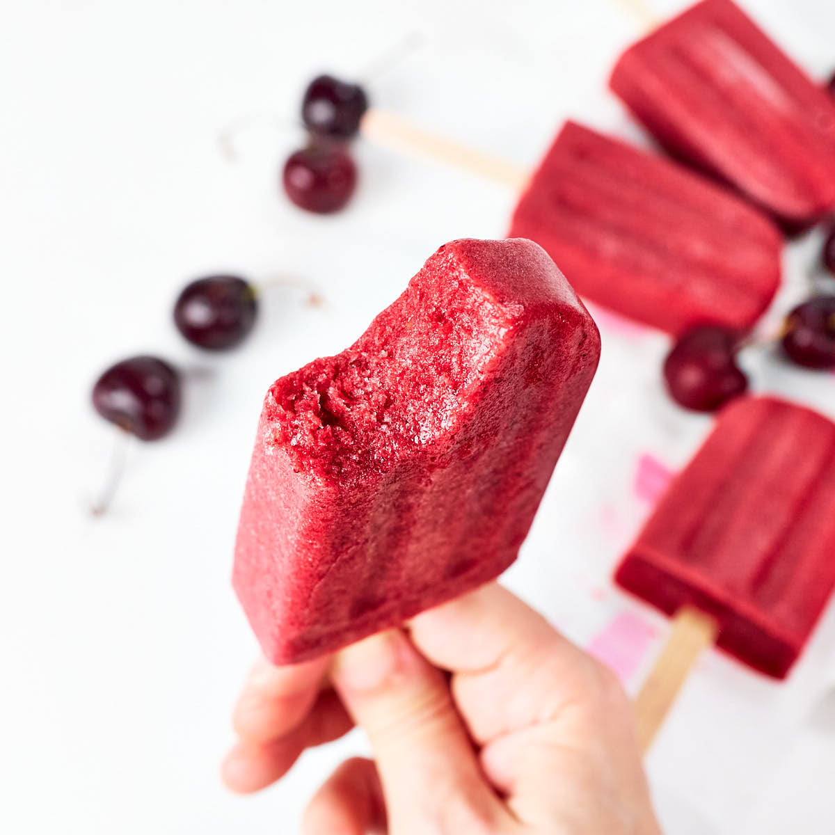 A hand holds a bitten cherry popsicle with more cherry popsicles and fresh cherries scattered in the background on a white surface. // FoodNouveau.com