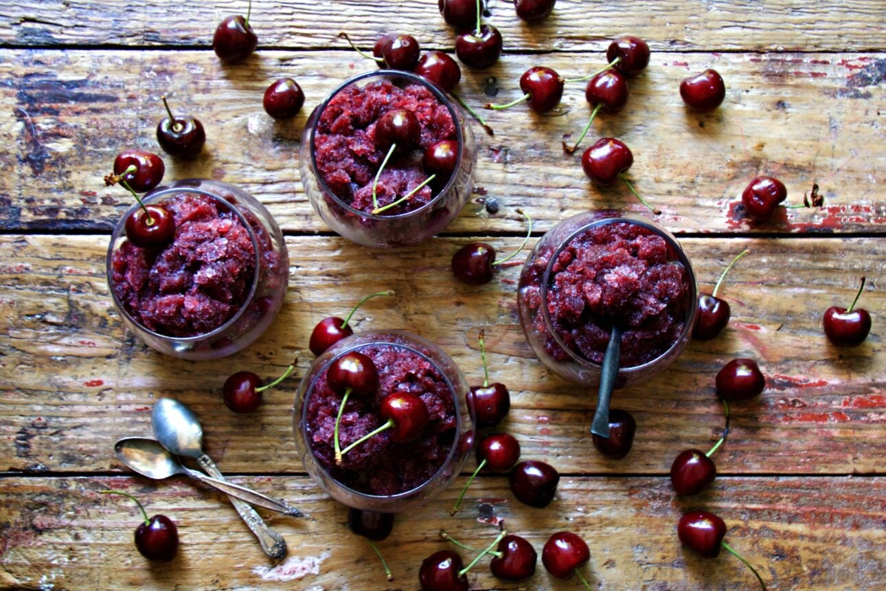 Four glass bowls of cherry granita on a rustic wooden table, surrounded by fresh cherries. Two spoons are placed to the side, and one bowl has a spoon inside. // FoodNouveau.com