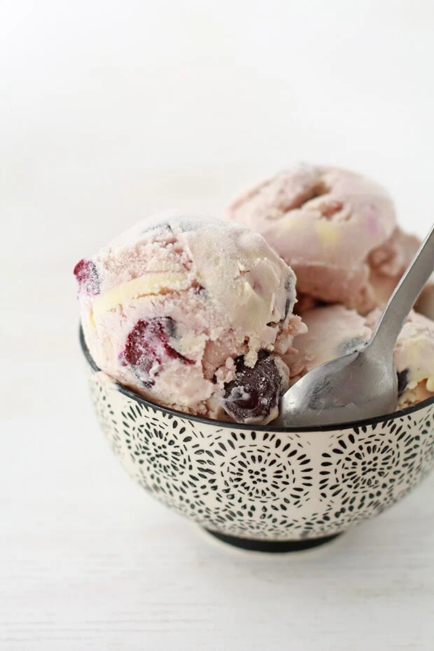 A patterned bowl filled with three scoops of cherry ice cream, visible cherry pieces, and a spoon resting inside on a white background. // FoodNouveau.com