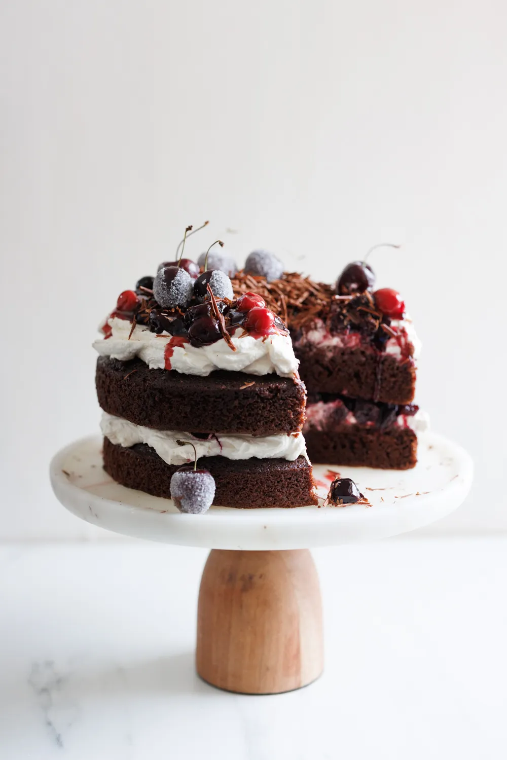 A chocolate layer cake with whipped cream, cherries, and chocolate shavings on top sits on a white cake stand; a large slice is missing. // FoodNouveau.com