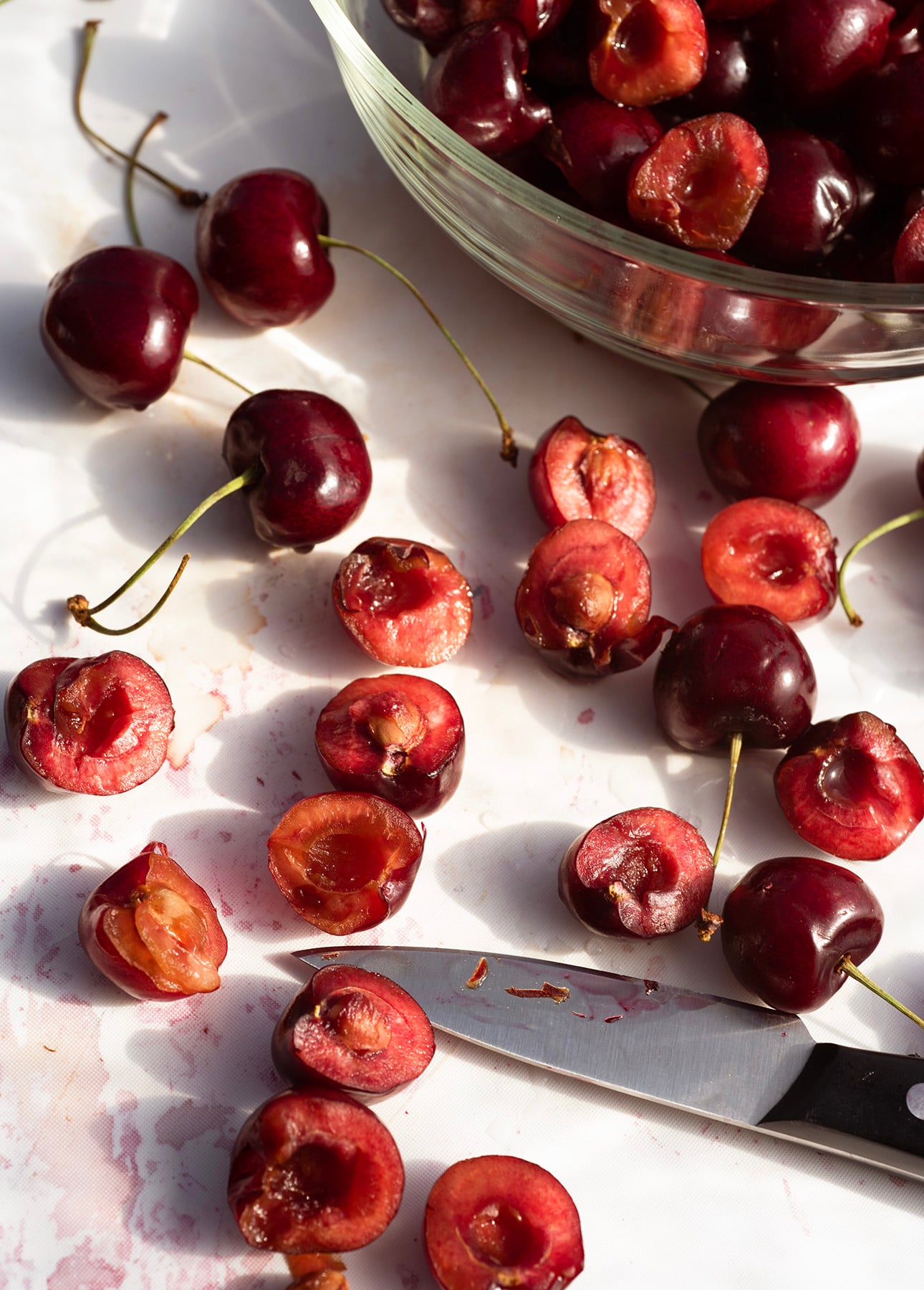 A bowl of whole cherries sits beside halved cherries and a knife on a white surface, with sunlight casting shadows across the scene—setting the stage for preparing a classic French cherry clafoutis. // FoodNouveau.com