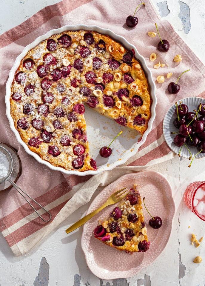 A cherry clafoutis in a round dish with a slice served on a pink plate. Fresh cherries, hazelnuts, and a gold fork are nearby on a striped cloth. The dessert is dusted with powdered sugar. // FoodNouveau.com