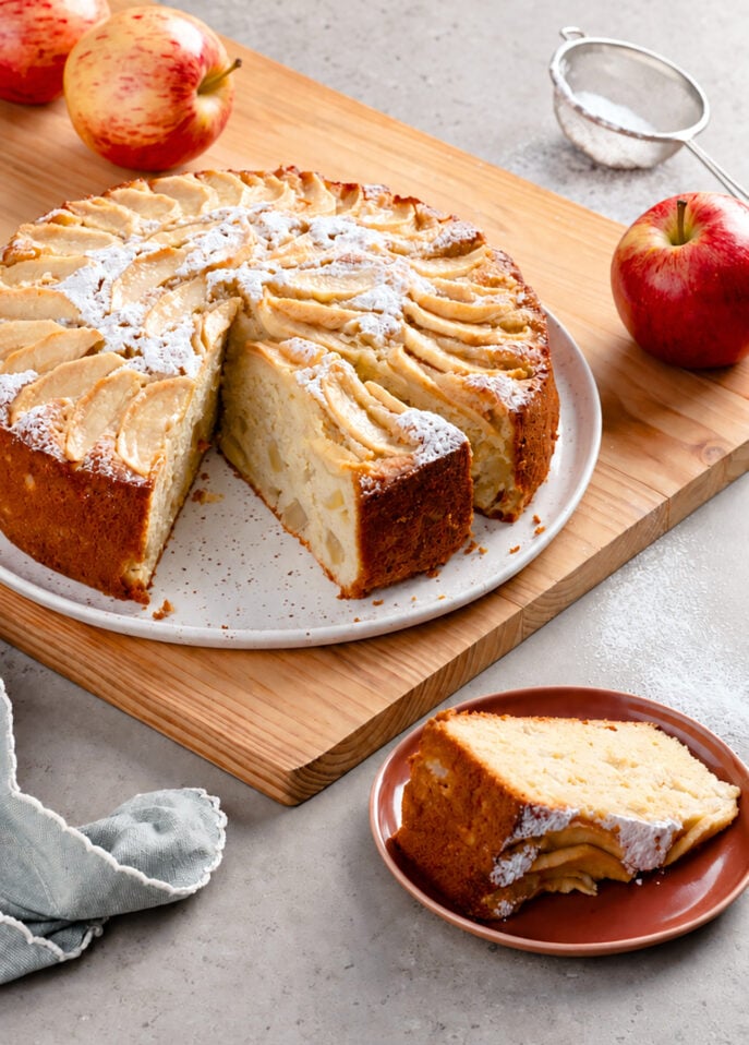 A sliced Italian apple cake topped with powdered sugar sits on a wooden board, with a single slice on a small plate and two whole apples nearby. // FoodNouveau.com